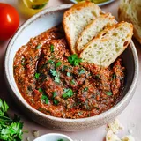 Eine Schüssel mit herzhaftem Tomatendip, garniert mit frischer Petersilie, dazu drei Scheiben Brot und einige Zutaten im Hintergrund.