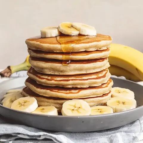 Ein Teller mit einem hohen Stapel von Pfannkuchen, garniert mit Bananenscheiben und Sirup, steht neben einer Banane.