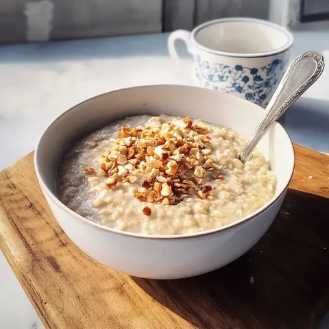 Eine Schüssel mit Haferbrei, garniert mit gehackten Nüssen, steht auf einem Holzbrett neben einer Tasse.