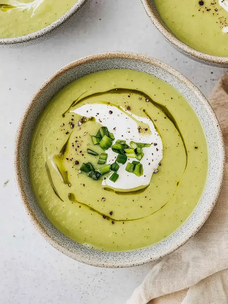 Eine Schüssel mit cremiger grüner Suppe, garniert mit Sauerrahm, frischen Schnittlauchstückchen und einem Spritzer Olivenöl.