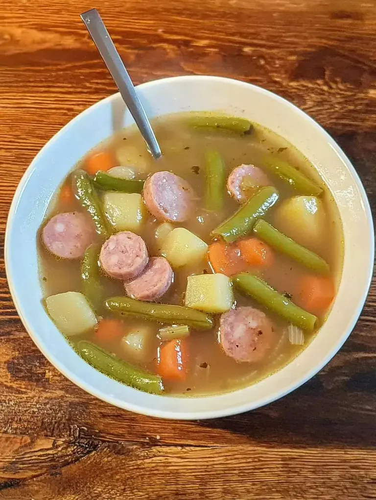 Eine Schüssel mit Gemüsesuppe, die Kartoffeln, Karotten, grüne Bohnen und Scheiben von Wurst enthält, steht auf einem Holztisch.
