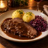 Ein Sauerbraten-Teller mit dunkler Sauce, Kartoffelklößen und Rotkohl in warmem Restaurantlicht.